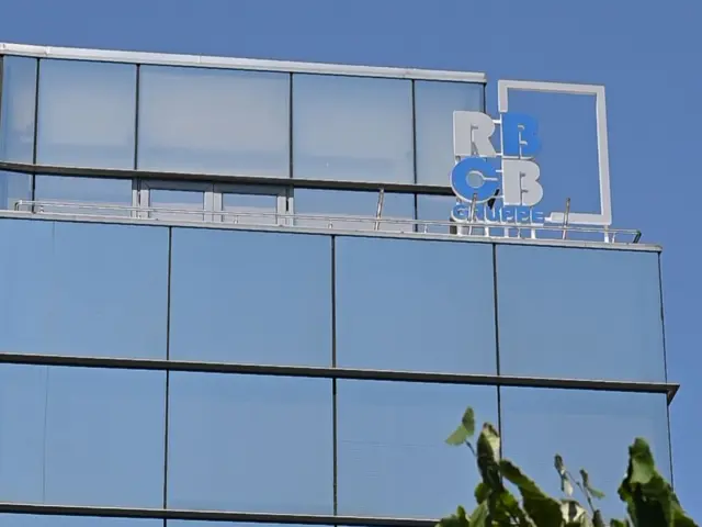Logo volumetric alb-albastru, încadrat geometric, montat la coronamentul unei clădiri cu fațadă vitrată.