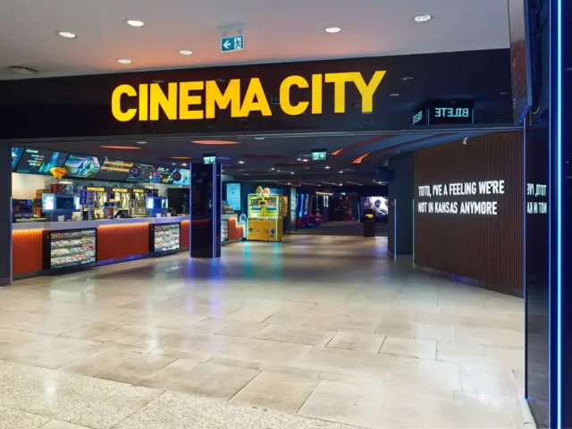 Litere volumetrice galbene CINEMA CITY deasupra accesului, cu zona de concesiuni vizibilă în interior.