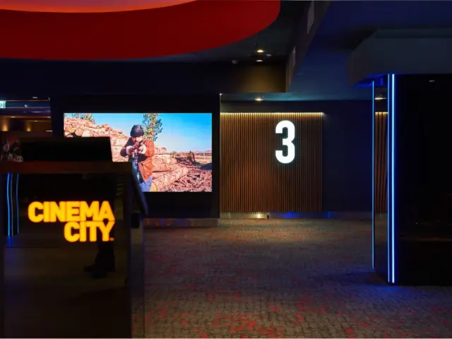Logo luminos Cinema City pe suport negru, lângă un ecran și intrarea în sala 3.