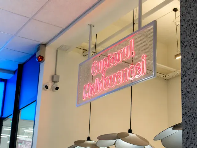 Semn Neon Flex roz „Cuptorul Moldovencei”, suspendat pe un panou tip plasă într-un spațiu interior.