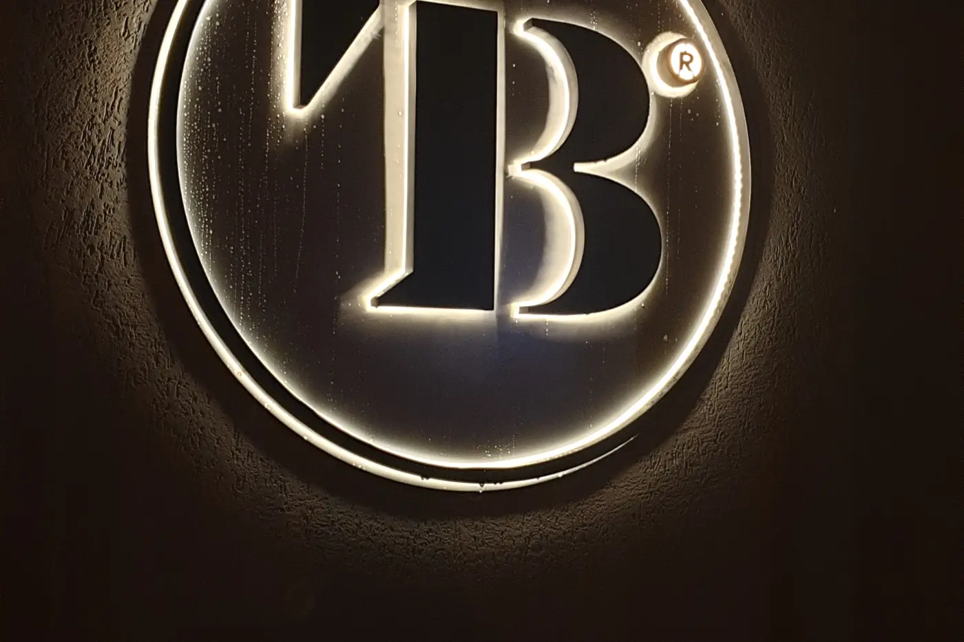 Monogram volumetric iluminat, încadrat într-o emblemă circulară montată pe un perete închis la culoare.