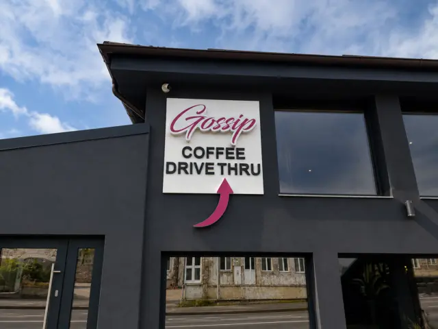 Panou Gossip Coffee Drive Thru cu litere volumetrice și săgeată roz, montat pe fațada închisă la culoare.