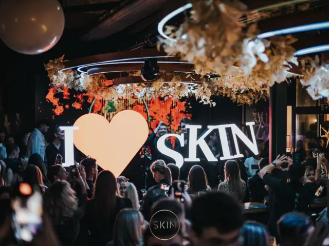 Ansamblu luminos „I ♥ SKIN”, cu litere albe și inimă centrală, expus într-un club aglomerat.