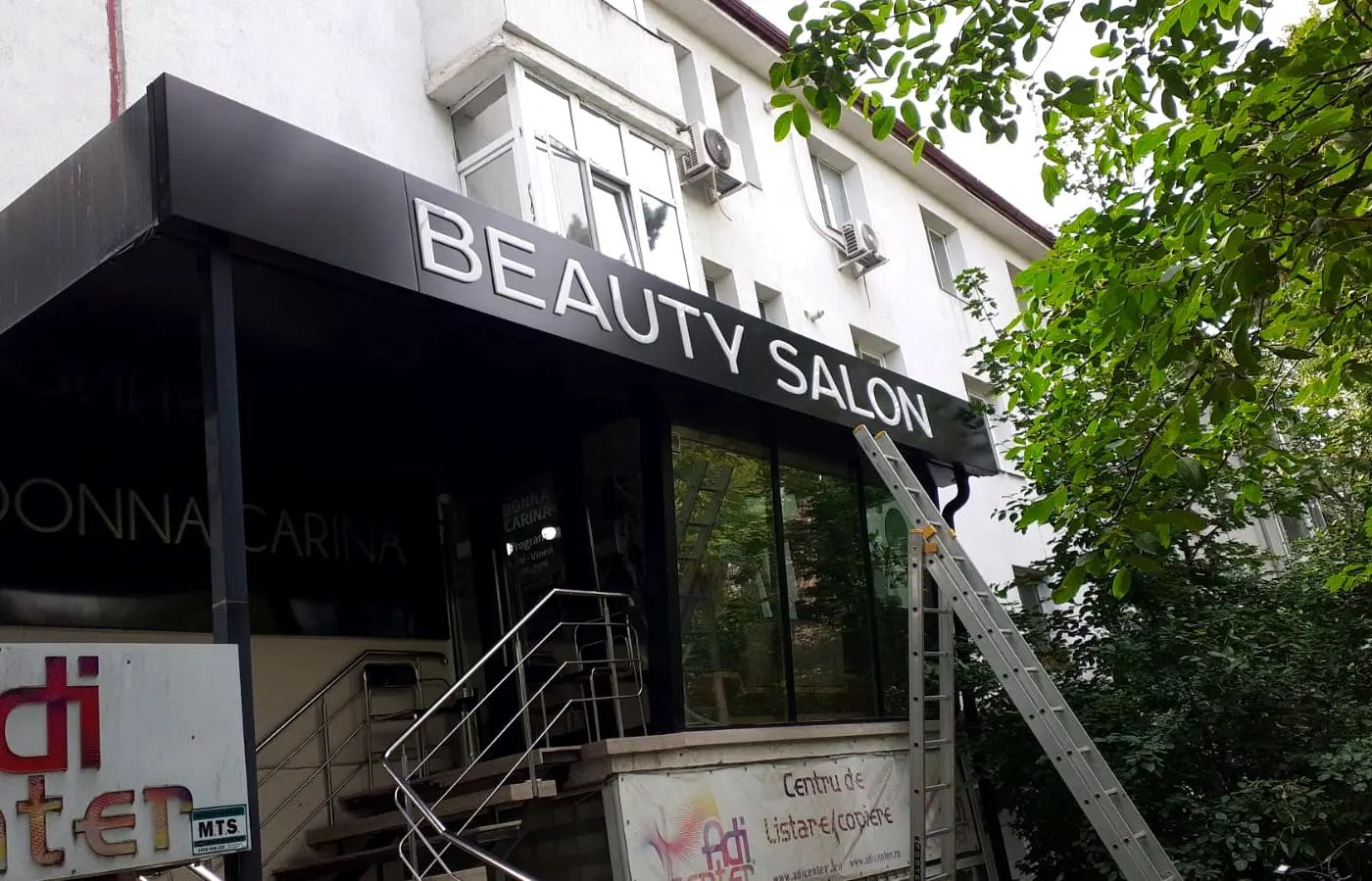 Litere volumetrice albe „BEAUTY SALON” pe o copertină neagră, cu o scară sprijinită de fațadă.