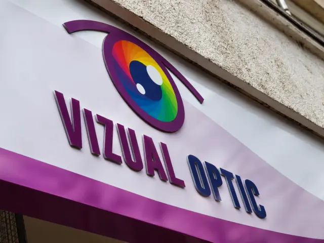 Siglă multicoloră și litere volumetrice Vizual Optic pe un panou alb de fațadă.