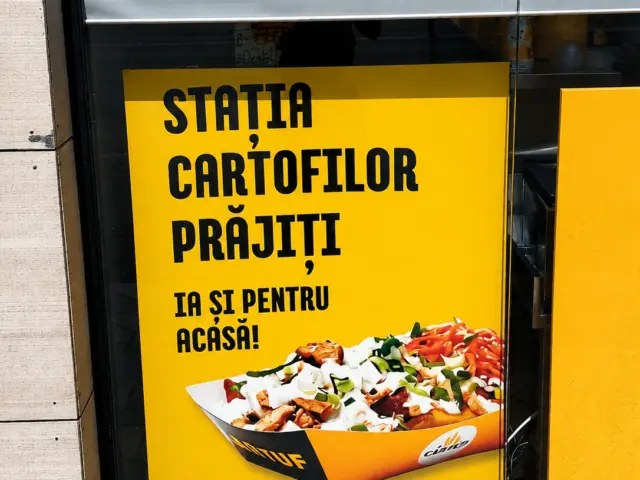 Panou luminos galben amplasat în vitrină, cu mesaj despre cartofi prăjiți și preparat ilustrat.