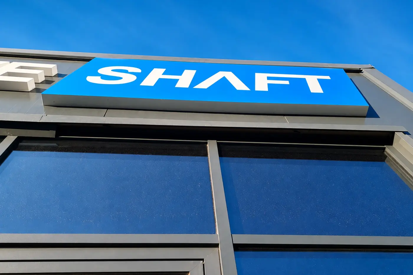 Casetă luminoasă albastră SHAFT, montată deasupra vitrinei unei clădiri cu fațadă gri.