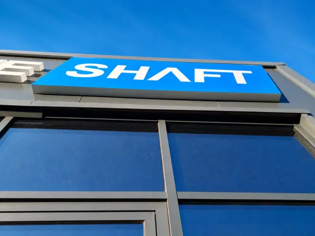 Blue SHAFT illuminated sign mounted above a glazed storefront on a gray facade.