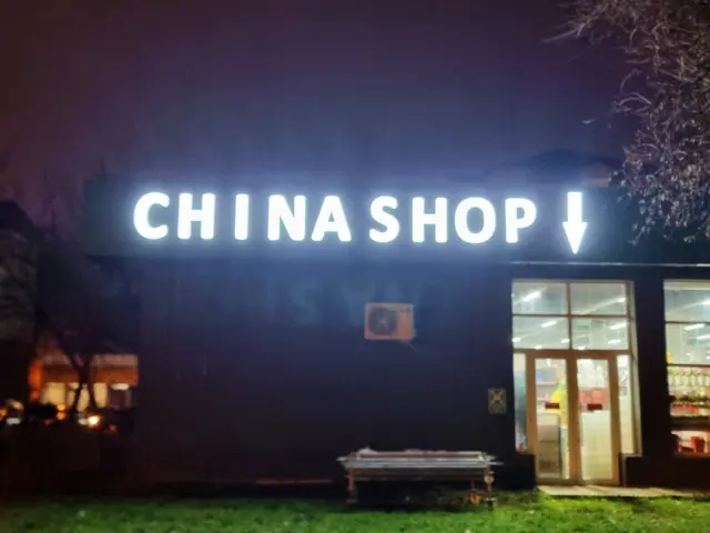 Literele luminoase CHINA SHOP și o săgeată în jos, montate pe fațadă deasupra intrării.