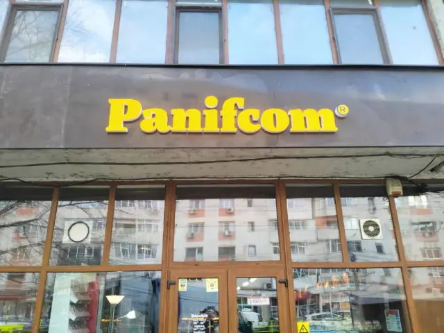 Litere volumetrice galbene Panifcom, montate pe panoul gri deasupra intrării vitrate.