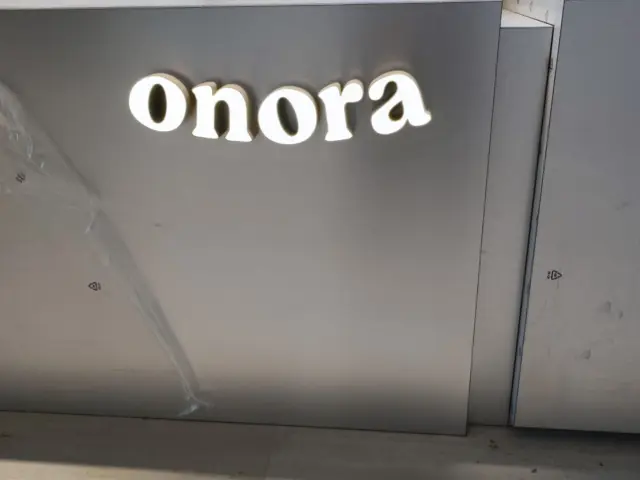 Literele luminoase Onora, aprinse pe un panou gri, într-un spațiu de lucru cu ambalaje.
