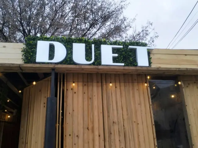 Litere volumetrice albe DUET, montate deasupra unei fațade placate cu lemn și a unui fundal vegetal.