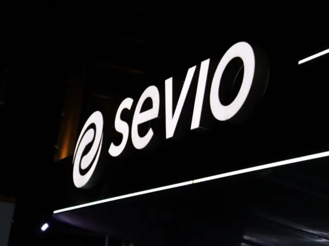 Logo-ul sevio cu litere volumetrice albe și emblemă circulară, iluminat pe o fațadă întunecată.