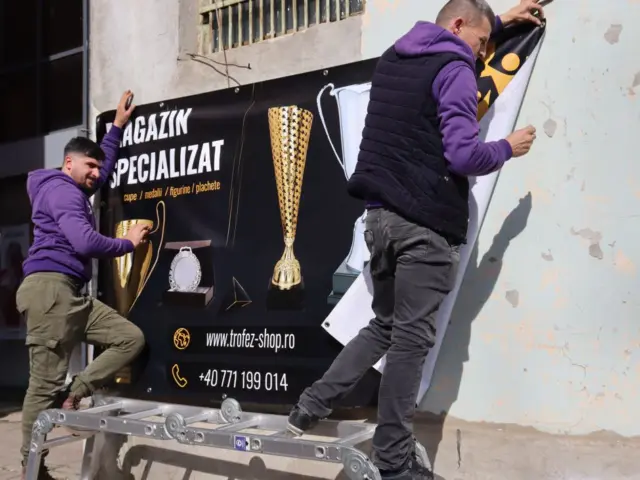 Doi tehnicieni montează pe fațadă bannerul Trofez Shop, decorat cu trofee, cupe și plachete.