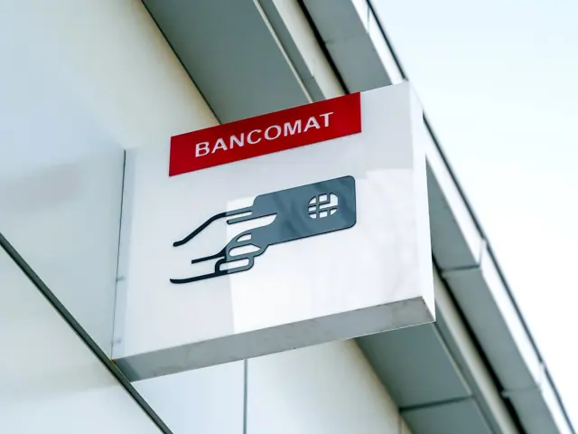 Semn perpendicular BANCOMAT, cu simbolul unei mâini ținând un card, montat pe fațada clădirii.