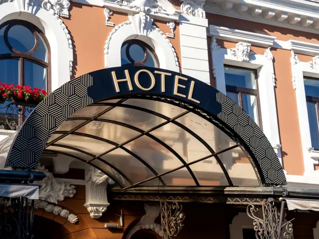 Litere volumetrice aurii „HOTEL”, montate pe o copertină arcuită, în fața unei clădiri cu fațadă ornamentată.