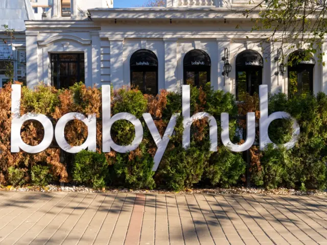 Litere volumetrice babyhub amplasate printre arbuști, în fața unei clădiri albe cu ferestre arcuite.