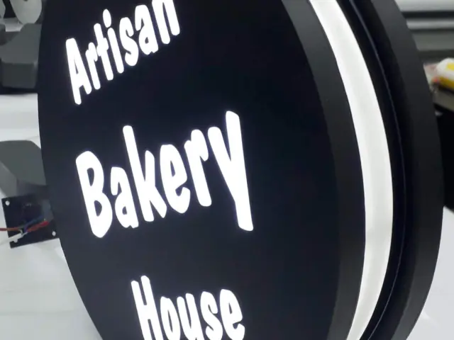 Firmă luminoasă rotundă, cu două fețe negre și text alb „Artisan Bakery House”, prezentată în atelier.