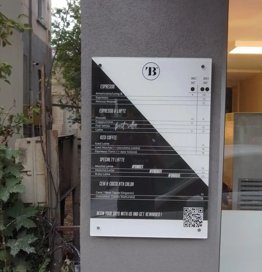 Meniu alb-negru pentru cafenea, cu prețuri și cod QR, montat pe peretele exterior lângă intrare.