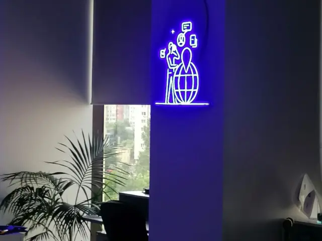 Semn Neon Flex alb și albastru, cu siluetă umană, glob și simboluri de călătorie, montat într-un birou.