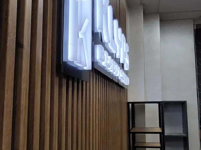 Litere și simbol volumetric iluminate, montate pe un panou închis la culoare, într-un interior cu lamele verticale.