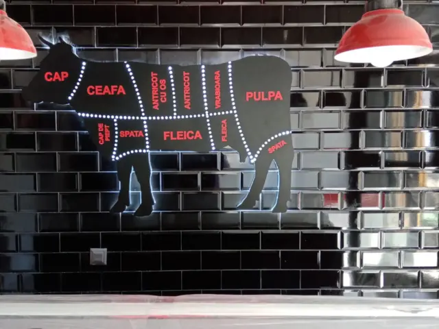 Siglă luminoasă fit beef și panou în formă de bovină, cu secțiuni de carne marcate, pe perete negru.