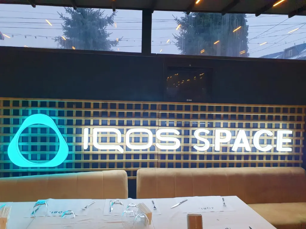 Firma IQOS SPACE, cu emblemă turcoaz și litere albe iluminate, montată pe un perete cu grilaj decorativ.