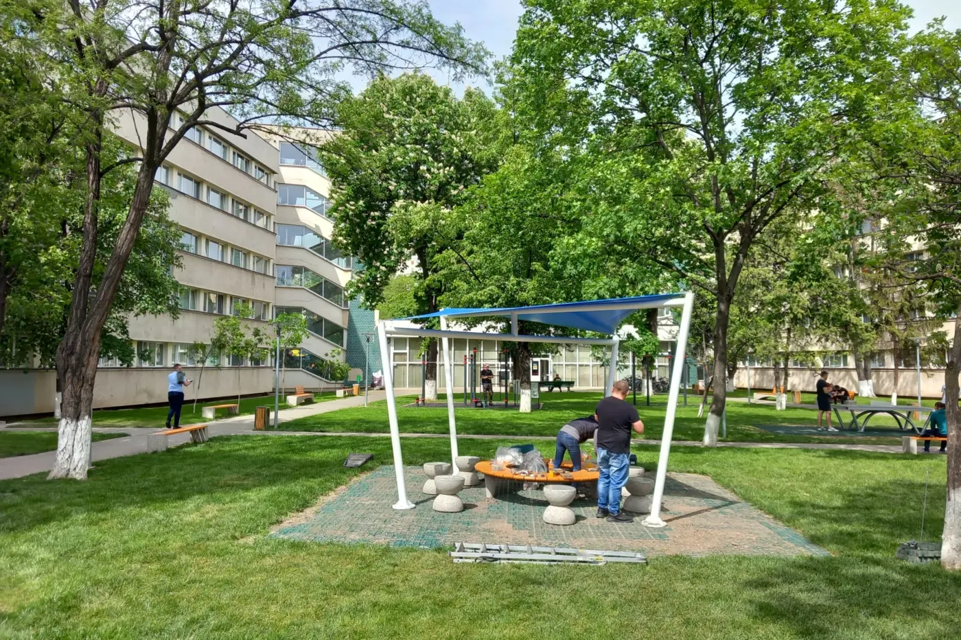 Structură metalică albă cu copertină albastră, în curs de montaj într-o curte verde.