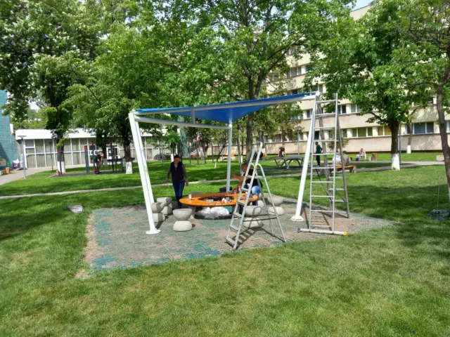 Structură metalică albă cu acoperire albastră, în curs de montaj într-un spațiu verde.