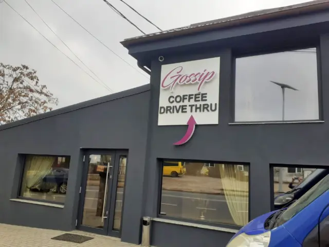 Semn volumetric Gossip Coffee Drive Thru, cu litere reliefate și săgeată roz, montat pe fațada gri.