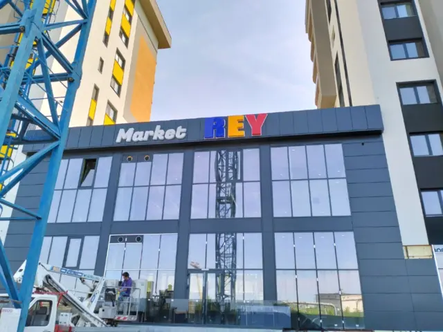 Litere volumetrice Market REY montate pe fațada vitrată, cu nacelă și structură metalică în prim-plan