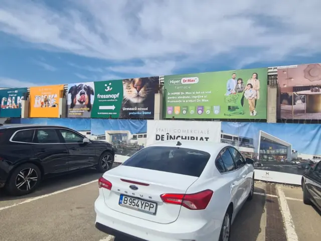 Ansamblu de bannere publicitare amplasate pe fațada unui centru comercial, deasupra parcării.
