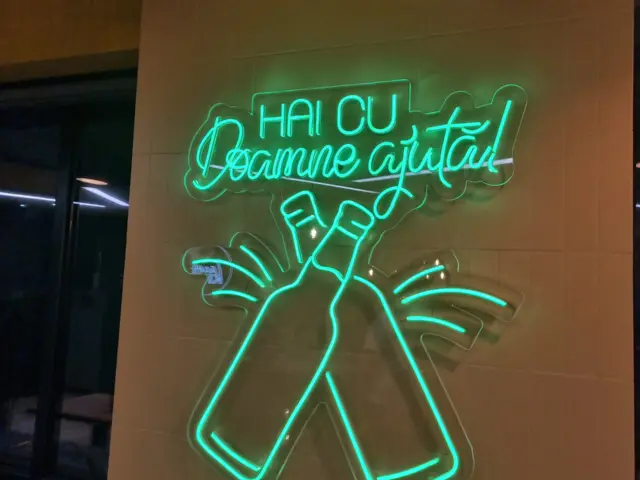 Semn verde Neon Flex cu mesajul „Hai cu Doamne ajută!” și două sticle încrucișate, montat pe perete.