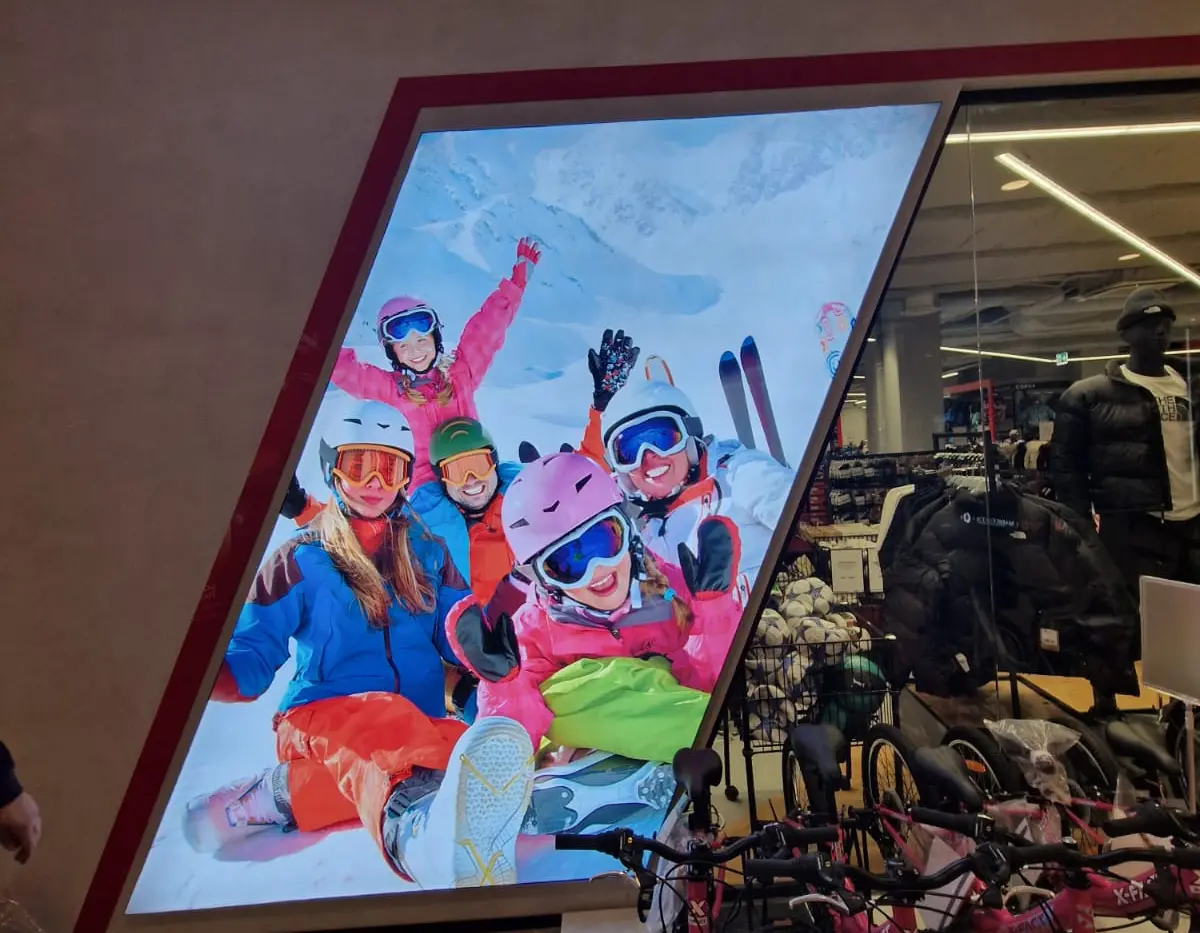 Panou publicitar luminos cu un grup de schiori, montat lângă intrarea unui magazin de articole sportive.