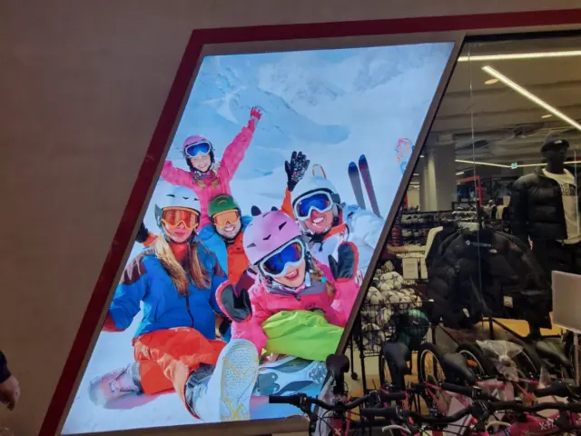 Panou publicitar luminos cu un grup de schiori, montat lângă intrarea unui magazin de articole sportive.