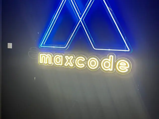 Sigla Maxcode din Neon Flex, cu simbol albastru și litere alb cald, montată pe un perete închis.
