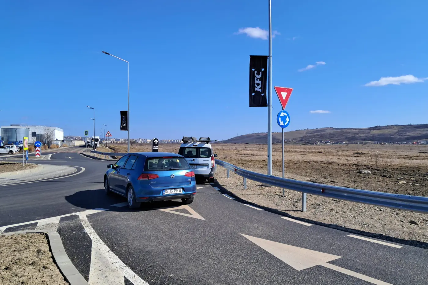 Banner negru KFC montat pe un stâlp stradal, lângă indicatoarele unui sens giratoriu.