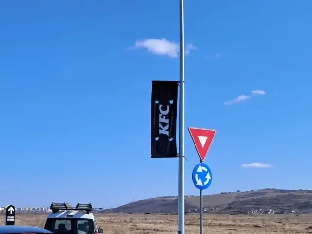 Banner negru KFC montat pe un stâlp stradal, lângă indicatoarele unui sens giratoriu.