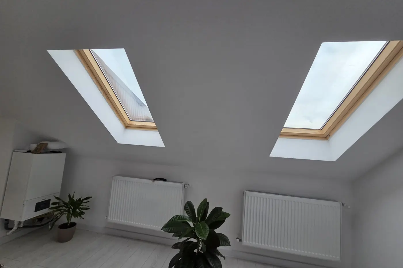 Două ferestre de mansardă cu folie de protecție solară, într-un interior alb cu radiatoare și plante.