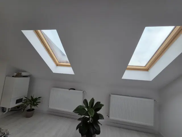 Două ferestre de mansardă cu folie de protecție solară, într-un interior alb cu radiatoare și plante.