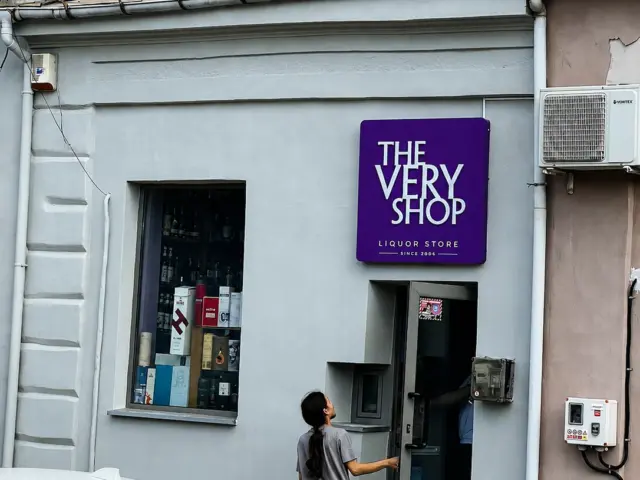 Firmă mov The Very Shop montată deasupra intrării, lângă vitrina cu sticle și cutii.