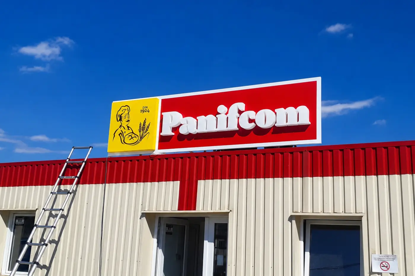 Firmă Panifcom cu litere albe volumetrice pe panou roșu, montată pe fațada unei clădiri industriale.