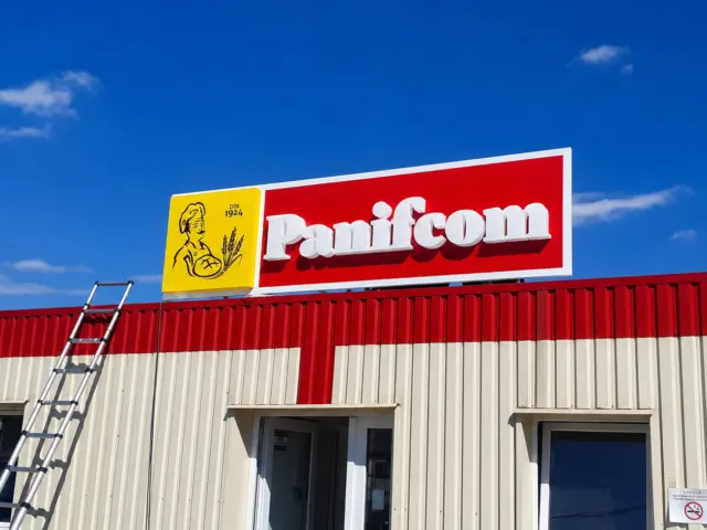 Firmă Panifcom cu litere albe volumetrice pe panou roșu, montată pe fațada unei clădiri industriale.