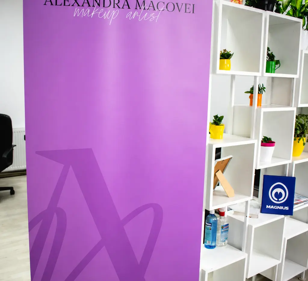 Roll-up mov Alexandra Macovei, makeup artist, expus într-un spațiu interior cu rafturi albe și plante.