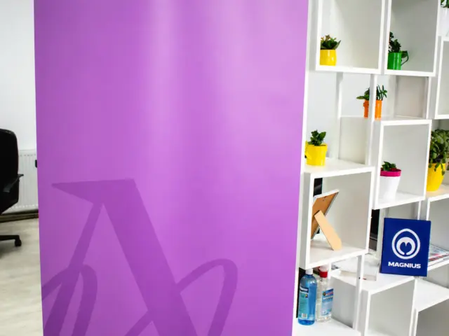 Roll-up mov Alexandra Macovei, makeup artist, expus într-un spațiu interior cu rafturi albe și plante.