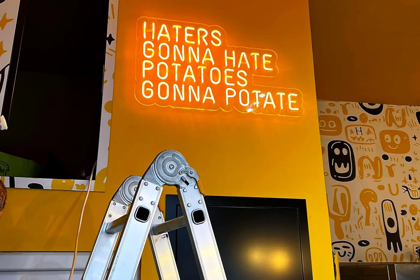 Semn Neon Flex portocaliu cu mesajul „Haters Gonna Hate, Potatoes Gonna Potate”, montat pe un perete galben.