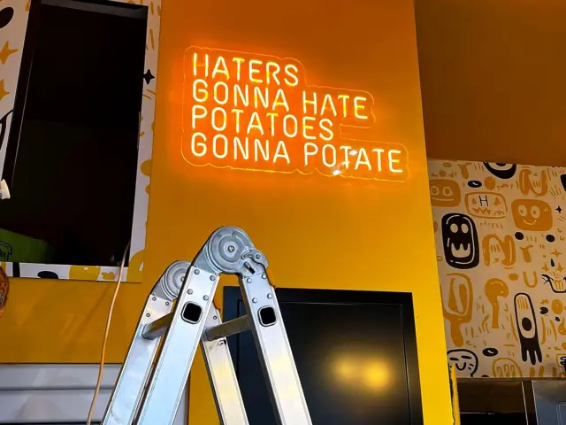 Semn Neon Flex portocaliu cu mesajul „Haters Gonna Hate, Potatoes Gonna Potate”, montat pe un perete galben.