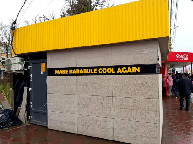 Litere galbene neluminoase „MAKE BARABULE COOL AGAIN”, montate pe o bandă neagră la exteriorul chioșcului.