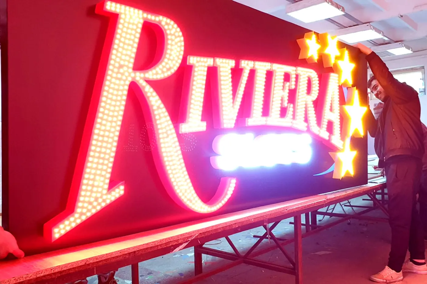 Reclamă Riviera cu litere și stele luminoase, manevrată de două persoane într-un atelier.