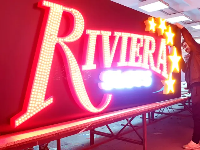 Reclamă Riviera cu litere și stele luminoase, manevrată de două persoane într-un atelier.